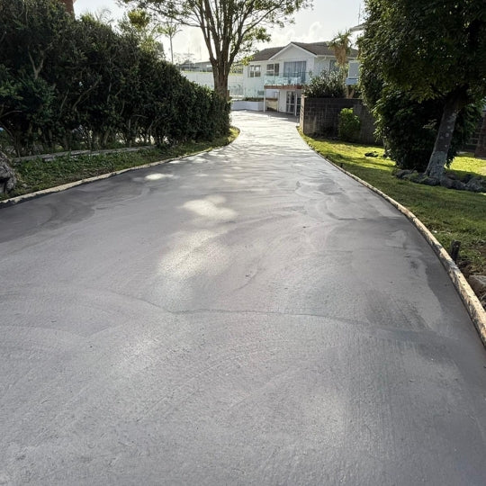 Wide charcoal concrete driveway curving up to a tree-screened Auckland home