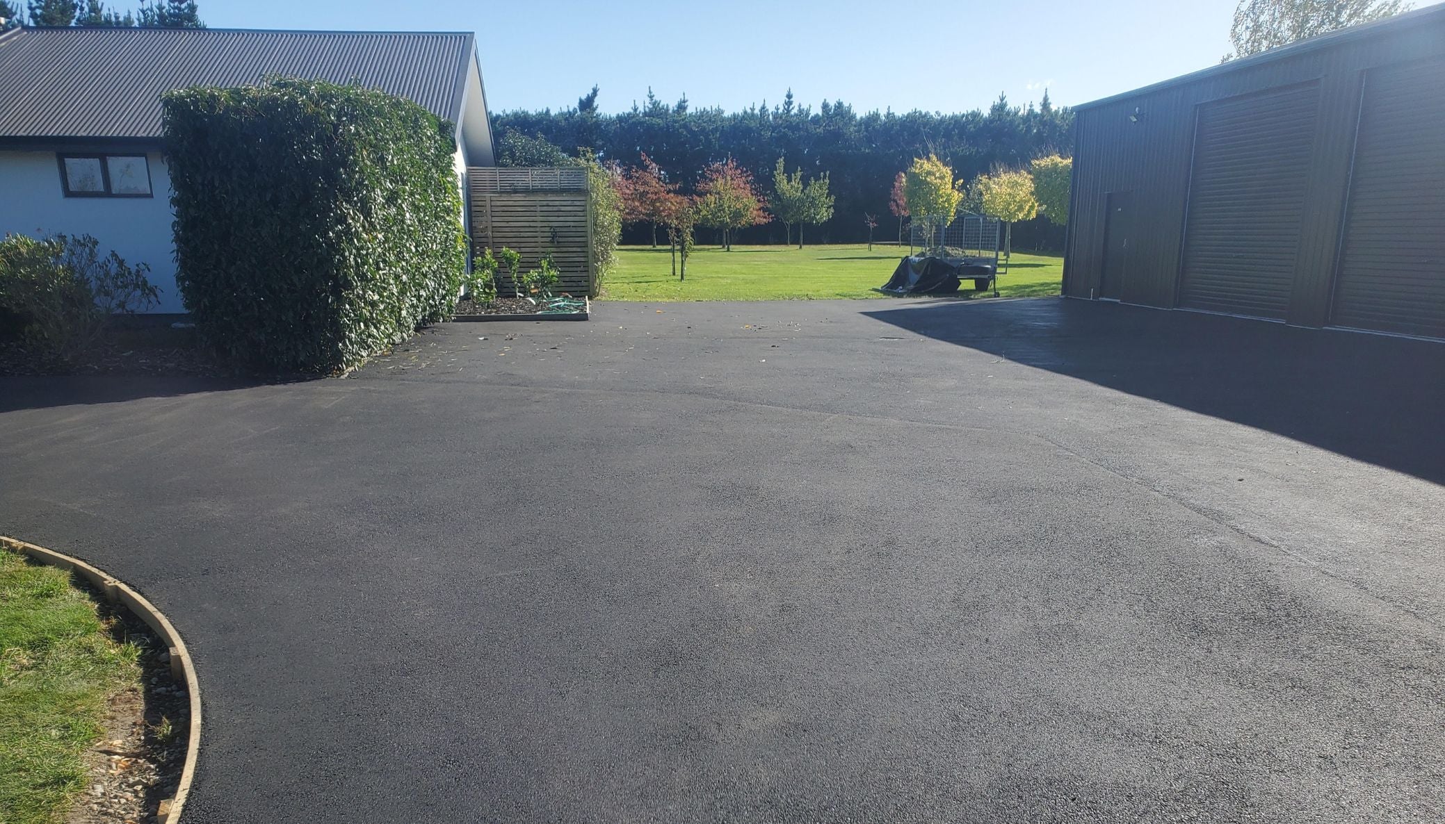 Wide asphalt driveway curving past a dark shed and green hedge